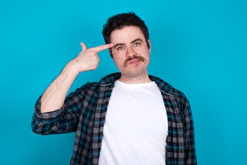 Wall Mural - Unhappy young Caucasian man with moustache wearing plaid shirt against blue wall curves lips and makes suicide gesture,  shoots in temple with hand, tries kill himself.