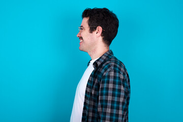 Wall Mural - Profile of smiling young Caucasian man with moustache wearing plaid shirt against blue wall with healthy skin, has contemplative expression, ready to have outdoor walk.