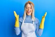 © Krakenimages.com - Beautiful blonde woman wearing cleaner apron and gloves celebrating mad and crazy for success with arms raised and closed eyes screaming excited. winner concept