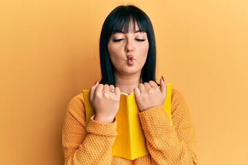 Wall Mural - Young hispanic woman holding book making fish face with mouth and squinting eyes, crazy and comical.