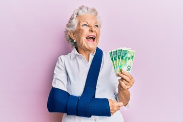 Wall Mural - Senior grey-haired woman wearing arm on sling holding israel shekels banknotes angry and mad screaming frustrated and furious, shouting with anger looking up.