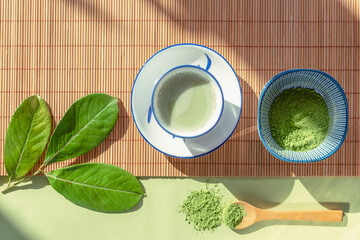 Green matcha tea in a white cup, tea powder and wooden spoon on bamboo napkin, top view. Healthy drink. Flat lay