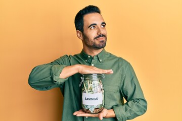 Poster - Young hispanic man holding jar with savings smiling looking to the side and staring away thinking.