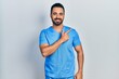 Handsome hispanic man with beard wearing blue male nurse uniform cheerful with a smile of face pointing with hand and finger up to the side with happy and natural expression on face