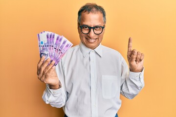 Sticker - Middle age indian man holding 100 philippine peso banknotes smiling with an idea or question pointing finger with happy face, number one