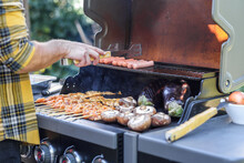 Close Up Shot Of An Anonymous Man Preparing Meat With Tongs On A Barbecue Outdoors. Hand Of Young Man Grilling Some Skewers, Meat Steak, Sausage And Vegetables On A Gas Grill, Bbq At Garden.