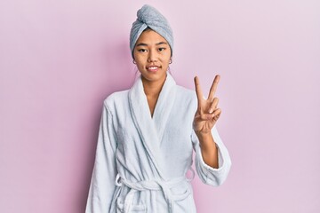 Poster - Young chinese woman wearing shower towel cap and bathrobe showing and pointing up with fingers number two while smiling confident and happy.