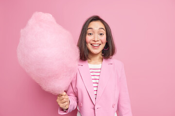 Wall Mural - Young cheerful young woman with eastern appearance holds big candy floss smiles joyfully being in good mood dressed in fashionable clothes Positive Asian female holds sugary dessert on stick