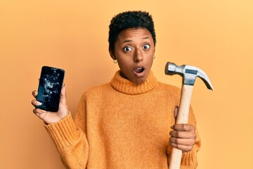 Wall Mural - Young african american girl holding broken smartphone showing cracked screen and hammer in shock face, looking skeptical and sarcastic, surprised with open mouth