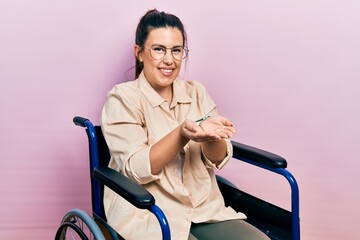 Poster - Young hispanic woman sitting on wheelchair smiling with hands palms together receiving or giving gesture. hold and protection