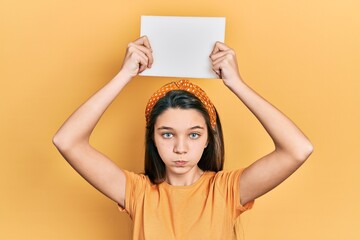 Poster - Young brunette girl holding blank empty banner over head puffing cheeks with funny face. mouth inflated with air, catching air.