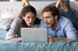 © fizkes - Stunned millennial couple relax in bed shocked by unexpected news on laptop. Amazed young man and woman lying in home bedroom look at computer screen surprised by message or notice on gadget.