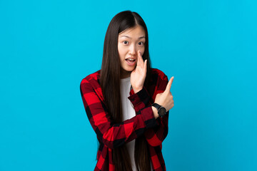 Wall Mural - Young Chinese girl over isolated blue background pointing to the side to present a product and whispering something