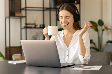 Online communication concept. Happy young caucasian millennial girl in headphones uses laptop, talking on video conference, gesturing hand sitting at the desk in the office