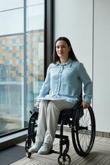 Wall Mural - Vertical full length portrait of young businesswoman in wheelchair smiling at camera while posing by window in office