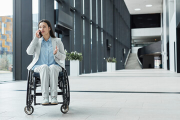 Wall Mural - Full length portrait of young businesswoman in wheelchair speaking by smartphone in office lobby