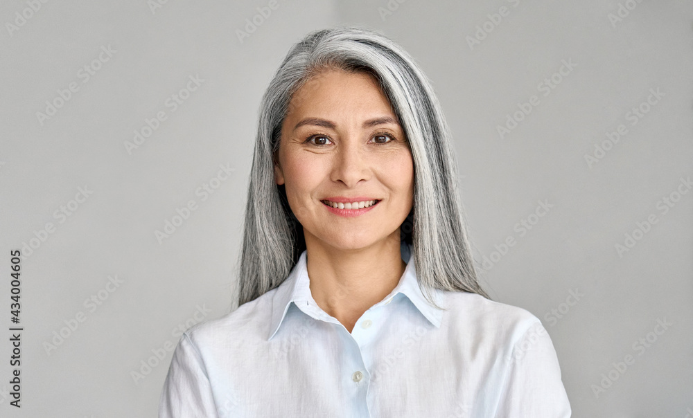 Happy smiling stylish confident 50 years old Asian female professional standing looking at camera at gray background. Portrait of sophisticated grey hair woman advertising products and services. - obrazy, fototapety, plakaty 