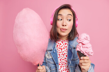 Wall Mural - Dark haired woman looks at delicious ice cream licks lips holds cotton candy enjoys eating summer dessert listens audio track via headphones dressed in fashionable clothes isolated over pink wall