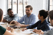 © fizkes - Satisfied millennial employee sharing ideas with diverse team on office brainstorming meeting, speaking and smiling. Mentor training multiethnic group of interns, talking to new hired about job tasks
