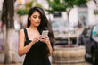 Young woman texting a silver smartphone while wearing a black dress and standing outside on the street