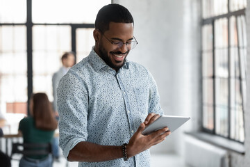 Happy Black male employee reading on digital device screen, using on tablet pc in modern office. African American manager shopping on internet, making video call. Developer testing online app