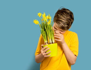 Wall Mural - Smiling caucasian shy teen boy hiding behind pot of daffodils in his hand. Happy Mother's or Teachers day greeting card on blue. Copy space
