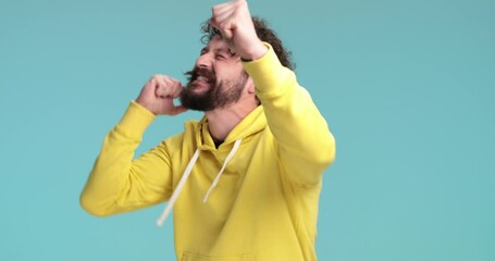 Wall Mural - shocked cool hipster man with long beard and moustache making a surprised face, holding fists in the air, dancing and celebrating victory while yelling and having fun on blue background in studio