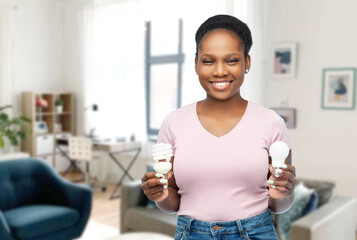 Poster - eco living, inspiration and sustainability concept - portrait of happy smiling young african american woman comparing lighting bulbs over home room background