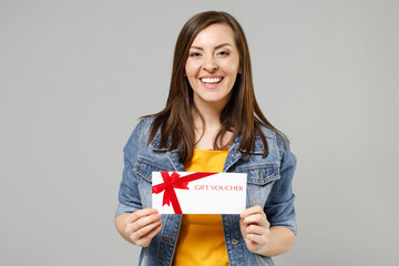 Wall Mural - Young smiling happy caucasian woman in casual trendy denim jacket yellow t-shirt holding in hand gift voucher flyer mock up look camera isolated on grey background studio. People lifestyle concept