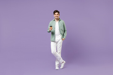 Full length young satisfied smiling fun man 20s wearing casual green mint shirt white t-shirt holding paper cup of coffee drink hot tea in morning isolated on purple color background studio portrait.