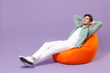 Full length fun man 20s in casual mint shirt white t-shirt sitting in orange bean bag chair hold hands folded behind neck isolated on purple color background studio portrait People lifestyle concept.