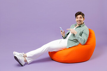 Full length man 20s wearing casual mint shirt white t-shirt sitting in orange bean bag chair point finger on mobile cell phone chat isolated on purple color background studio People lifestyle concept.