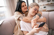 © LIGHTFIELD STUDIOS - lesbian couple hugging while sitting on couch and using smartphone