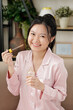 © DragonImages - Portrait of cheerful young woman in pink silk pajamas applying serum with hyaluronic acid on her face