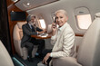 © LIGHTFIELD STUDIOS - Smiling businesswoman talking on smartphone and looking at camera near blurred businessman in plane