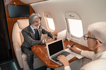 Businessman using laptop near blurred businesswoman writing on paper and champagne in private jet