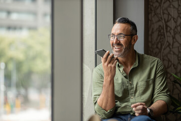 wide shot mature man with beard talking on phone next to sunny window