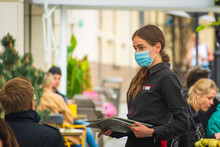 Pretty Nice Waitress With Mask And Menu Serving Clients At The Table Of An Outdoor Bar, Cafe Or Restaurant, Reopening After Lockdown And Restrictions 