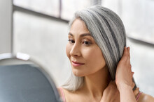 Portrait Of Beautyful Happy Middle Aged Mature Asian Woman, Senior Older 50s Lady Pampering Touching Gray Hair Looking At Herself At Mirror Indoors. Ads Of Lifting Anti Wrinkle Skin Hair Care Spa.