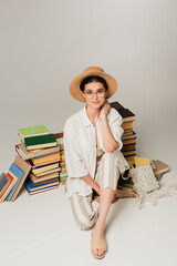 Wall Mural - full length of pleased woman in sun hat and glasses sitting near pile of books on white