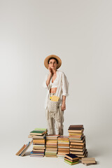 Wall Mural - pleased young woman in sun hat standing near pile of books isolated on white