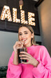 © Julio Rodriguez/ADDICTIVE STOCK - Cheerful young female in pink sweater sipping cold fizzy soft drink through straw while spending free time in cafeteria and looking at camera happily