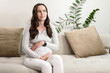 © Руслан Галиуллин - pregnant puzzled woman in gray blouse sits on couch thinking