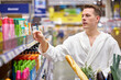 © Roman - Side view on young man need new shampoo or shower gel, came in supermarket in bathrobe before getting shower, choosing shampoo at discount