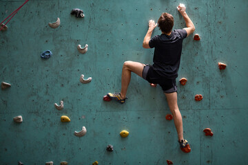 active young caucasian man on rock wall in sport center, rock climbing. Sportive male guy in black sportswear engaged in sport, fitness, lead healthy lifestyle. Rear view