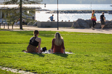 Two young women sit on the green grass near the city embankment. Relaxation by the lake on a warm sunny day