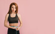 © Daria Lukoiko - Fitness girl smiles and measures the waist in black sportswear on a pink background. Slim woman with a beautiful athletic body and tanned skin