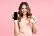 © GVS - Smiling pretty female pointing finger at her mobile phone and looking to camera on pink background