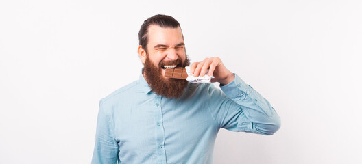 Portrait of bearded man in casual eating bar of chocolate over white background