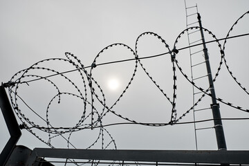 Barbed wire over a metal wall with a grid and a tower with gray sky and dim sun